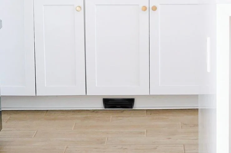 A black VacPort set into the toe kick under white kitchen cabinets, flush with a wood floor